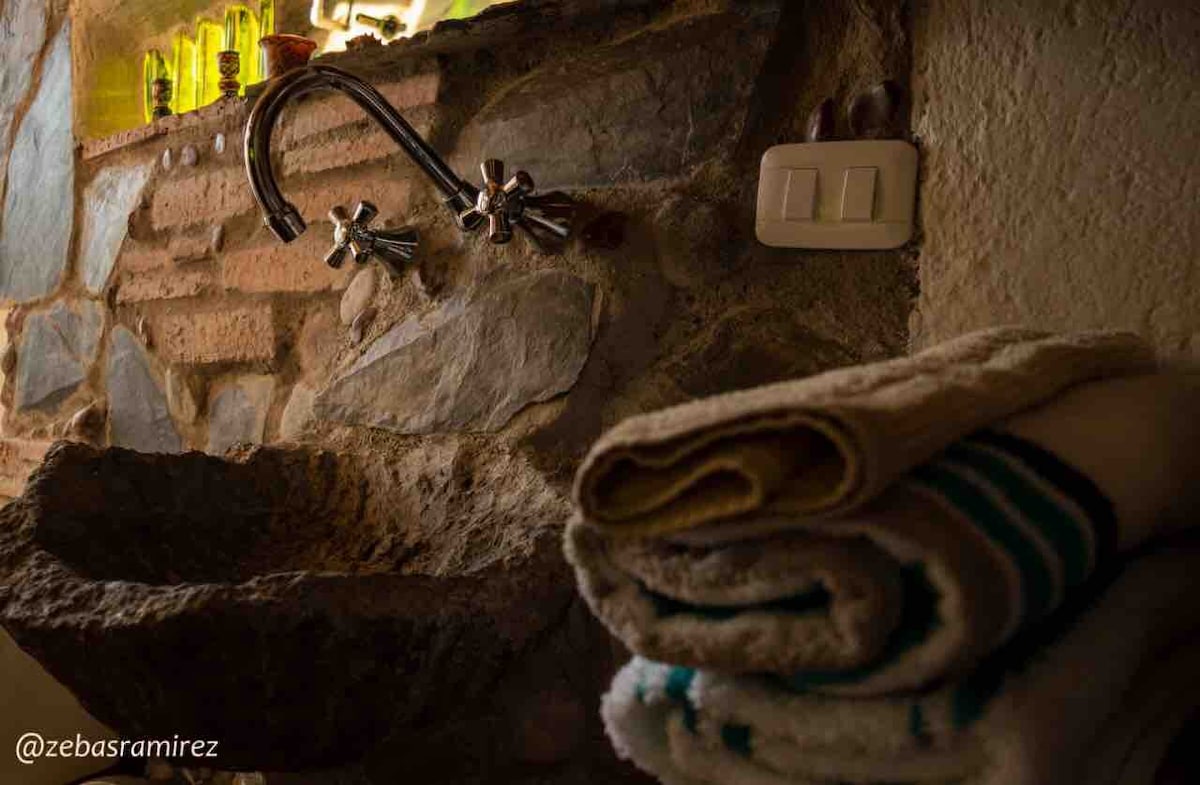 A stone sink with vintage-style faucets is positioned against a raw stone wall. Soft, neatly folded towels in various colors are stacked beside the sink, offering a functional and rustic touch to the bathroom area.