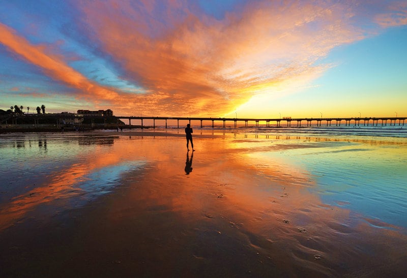 A tranquil beach scene is captured at sunset, with vibrant colors reflecting off the wet sand. A silhouette of an individual stands along the shoreline, while the pier stretches across the background, framed by colorful clouds in the sky.