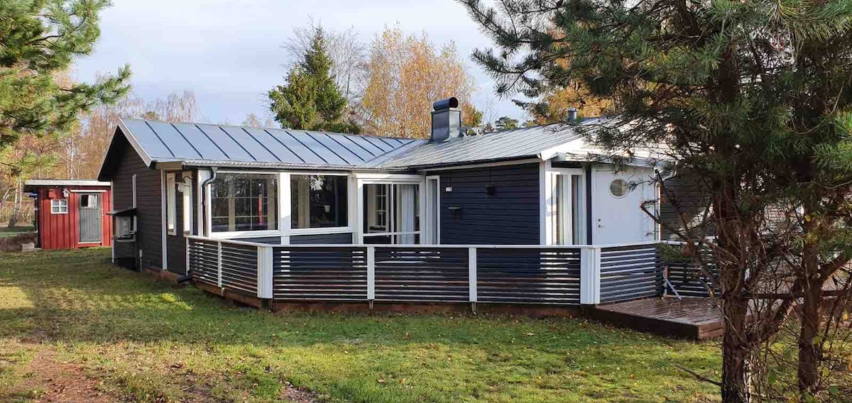 A single-story house features dark blue siding and a gray metal roof. Large glass doors provide access to a spacious deck surrounded by a white railing. Trees and greenery are present in the yard, creating a serene outdoor setting.