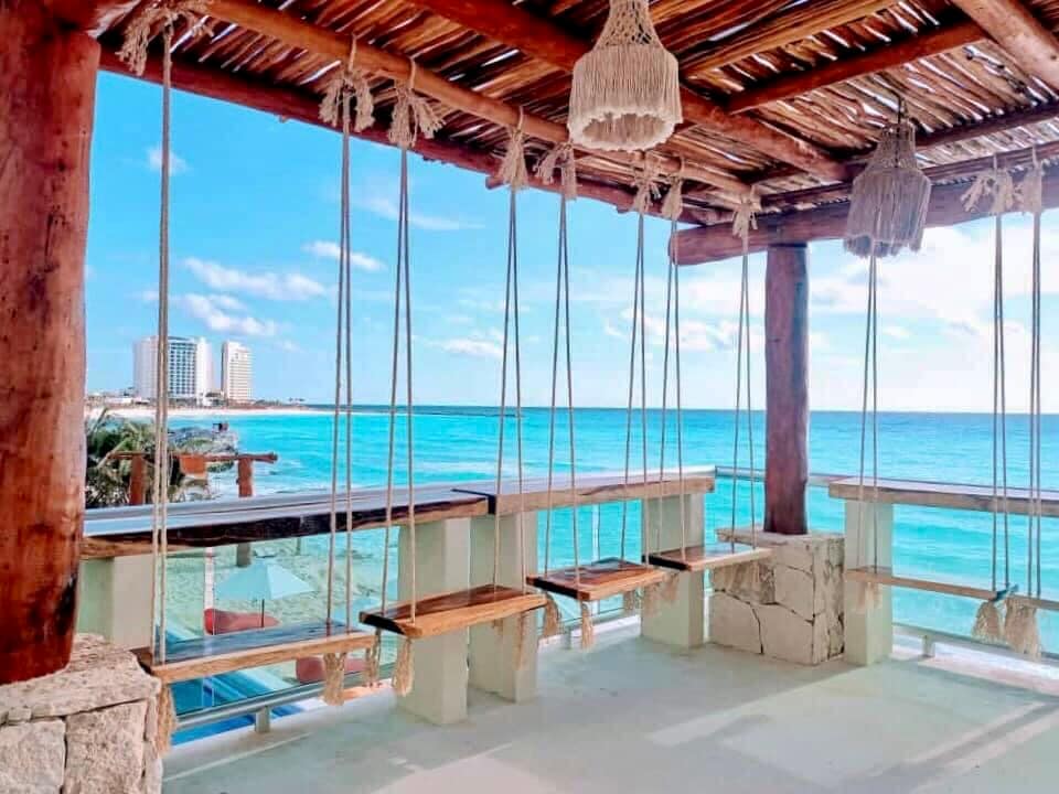 A spacious outdoor seating area features swings hanging from a wooden pergola, overlooking the ocean. The inviting turquoise water and beach can be seen in the background, complemented by a clear blue sky, creating a relaxing ambiance suitable for leisurely moments.