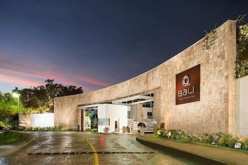 The entrance to the Bali Villas & Sports Club features a modern architectural design with a warm stone facade. A circular driveway allows for easy access, framed by lush greenery and ambient lighting as the sun sets in the background.