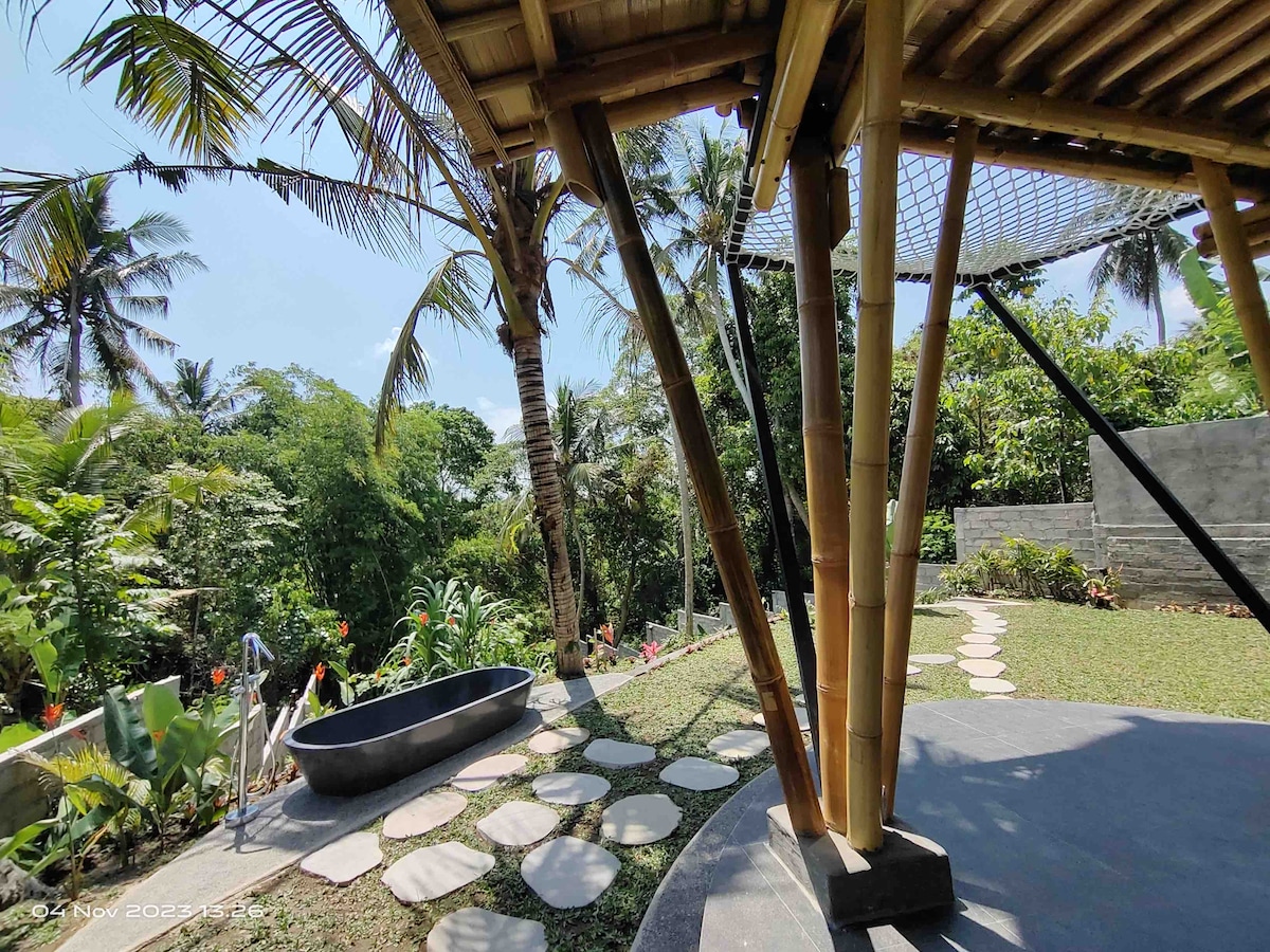An outdoor view of a bamboo structure is framed by palm trees and lush vegetation. A pathway of stepping stones leads to a modern bathtub, surrounded by vibrant landscape features. Natural light brightens the serene setting, emphasizing the tranquil atmosphere.