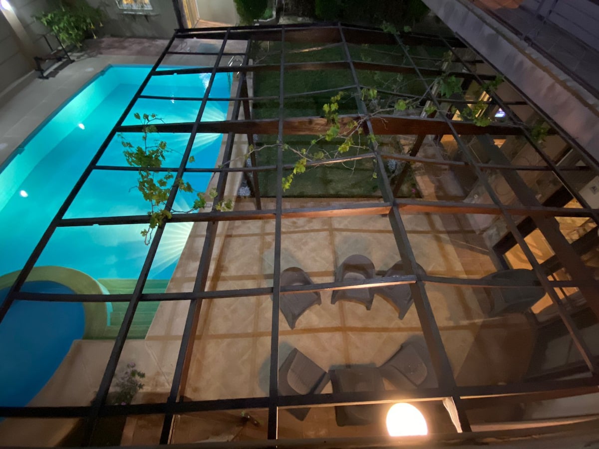 An aerial view captures the private pool and garden area. The pool shines in blue, surrounded by a spacious patio featuring a circular seating arrangement with several chairs. Greenery from the surrounding plants adds a natural touch to the outdoor space.
