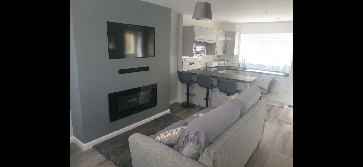 An open-plan living area is shown, featuring a three-seater sofa facing a mounted flat-screen TV. A modern kitchen with a sleek design includes bar seating for two. A soft rug is placed under the sofa, and natural light enters from large windows.