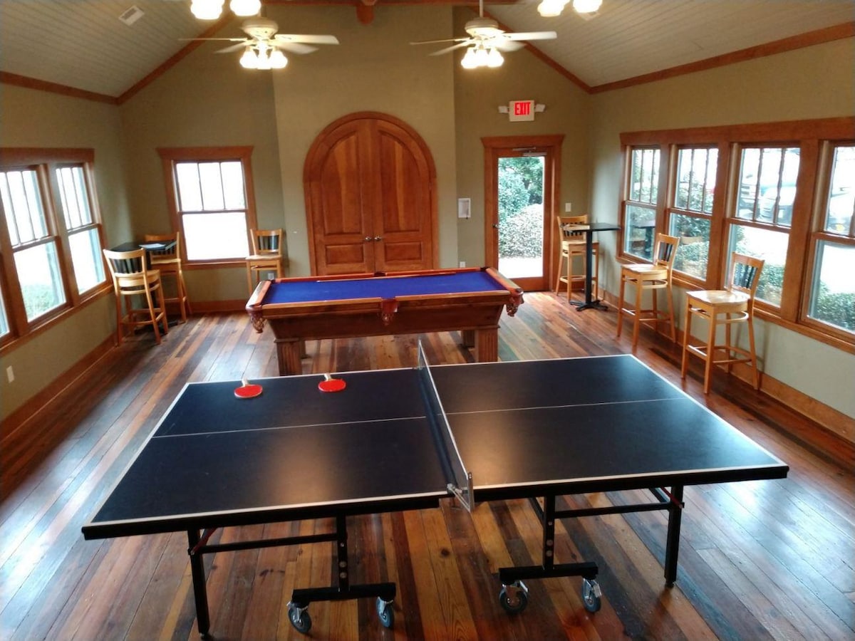 The game room features a ping pong table at the forefront and a pool table nearby. Large windows allow natural light to fill the space, highlighting the wooden flooring. Stools are arranged against the walls, providing additional seating options.