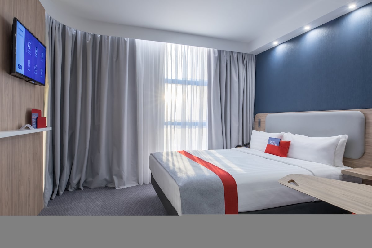 A modern guest room is highlighted by a spacious bed with crisp white linens and decorative pillows. Natural light streams through sheer curtains, illuminating the sleek grey walls and wooden accents. A flat-screen TV is mounted on the wall opposite.
