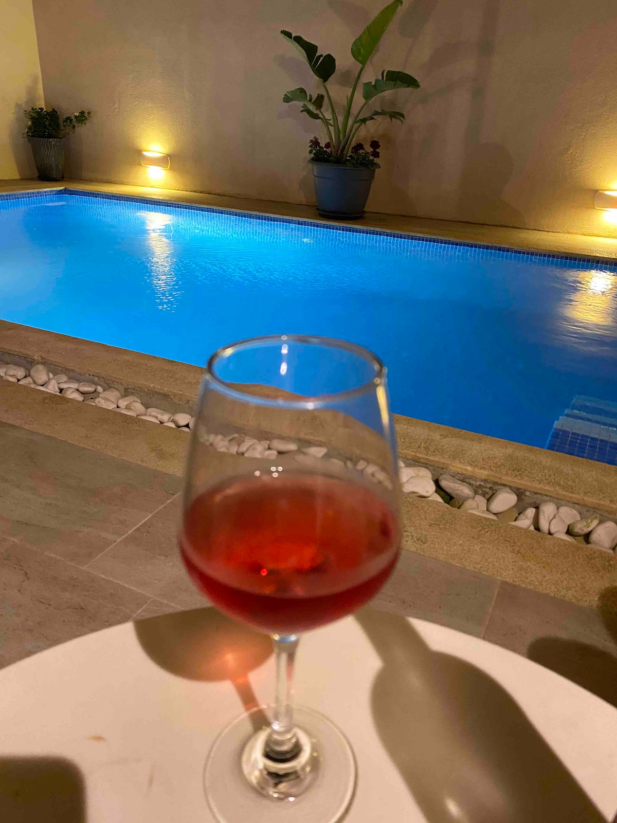 A glass of red beverage is set on a light-colored table in the foreground, with a softly lit swimming pool visible in the background. The pool's water reflects the warm illumination from the surrounding area, highlighting the calm and inviting atmosphere.