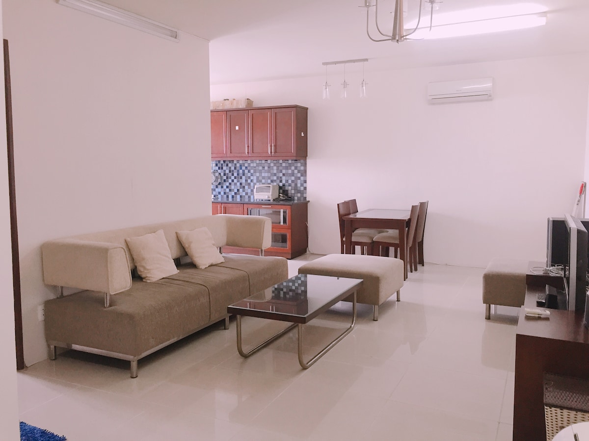 A spacious living area features a modern sofa with several cushions, a glass coffee table, and a cozy dining set. The kitchen, visible in the background, includes wooden cabinetry and essential appliances, providing a functional space for meal preparation.