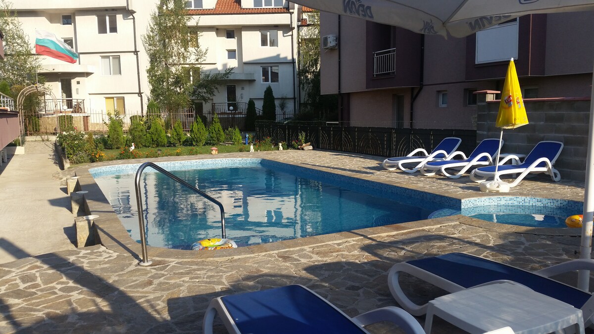 A rectangular swimming pool is surrounded by a stone patio, featuring several lounge chairs. Lush greenery and colorful plants are visible in the background, alongside residential buildings. An umbrella is positioned near the pool's edge, providing shade.