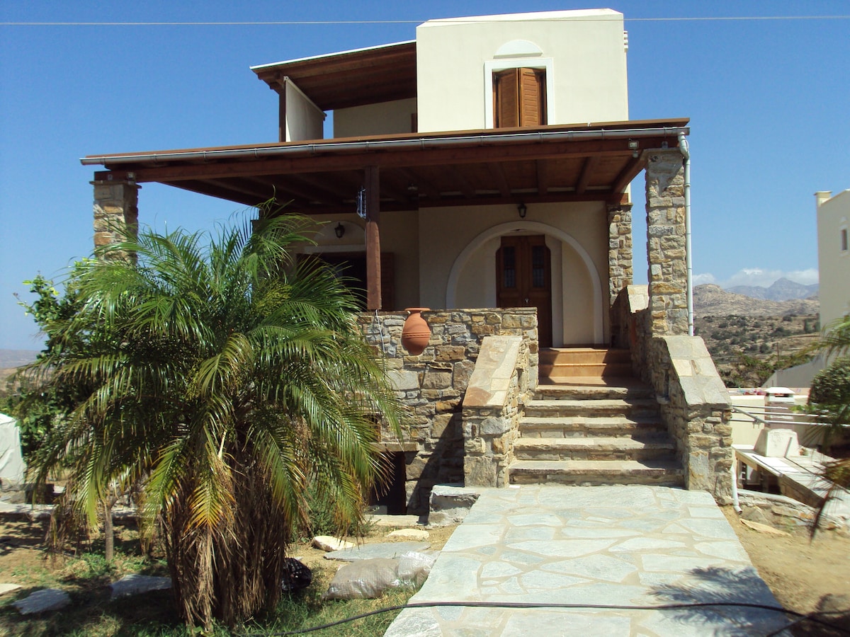 A two-story house is depicted, showcasing a mix of stone and plaster features. A spacious porch with wooden beams is highlighted, along with a staircase leading to the entrance. Lush greenery, including a palm tree, frames the pathway, enhancing the building's inviting appearance.