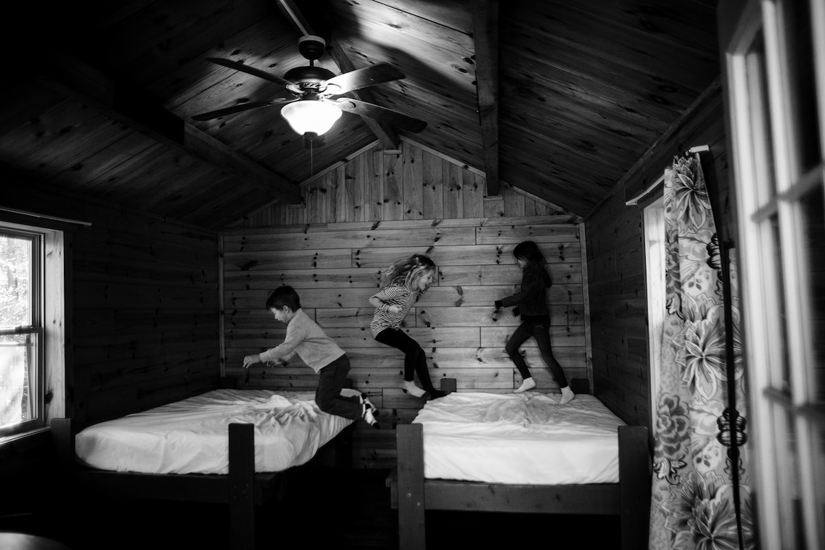 A rustic cabin interior is shown, featuring two wooden beds with white linens. Three children are depicted mid-jump on the beds, adding a sense of playfulness. Natural wood paneling and a ceiling fan contribute to the cozy atmosphere, with a window allowing soft light to enter.