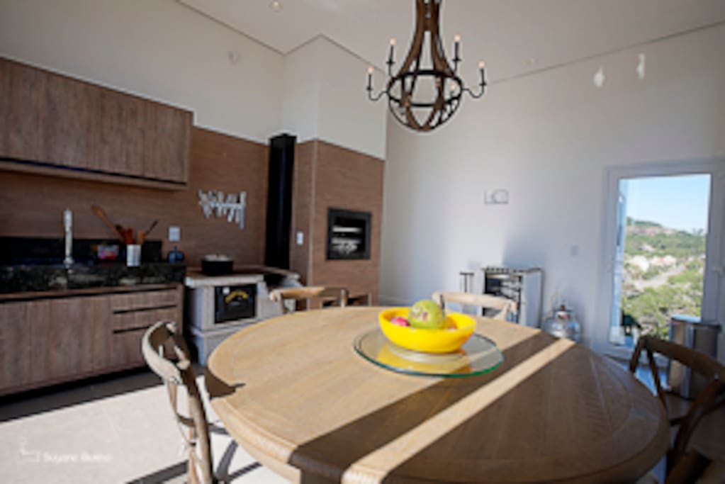 Segunda cozinha, mesa para 6 pessoas acomodadas. Com churrasqueira e saída para deck e piscina.