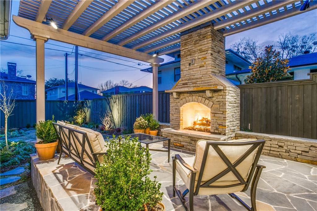 An outdoor patio area features two chairs positioned around a stone fireplace, surrounded by decorative plants. The space is partially covered by a pergola, offering a cozy setting for relaxation. Warm light glows from the fireplace, enhancing the inviting atmosphere during twilight.