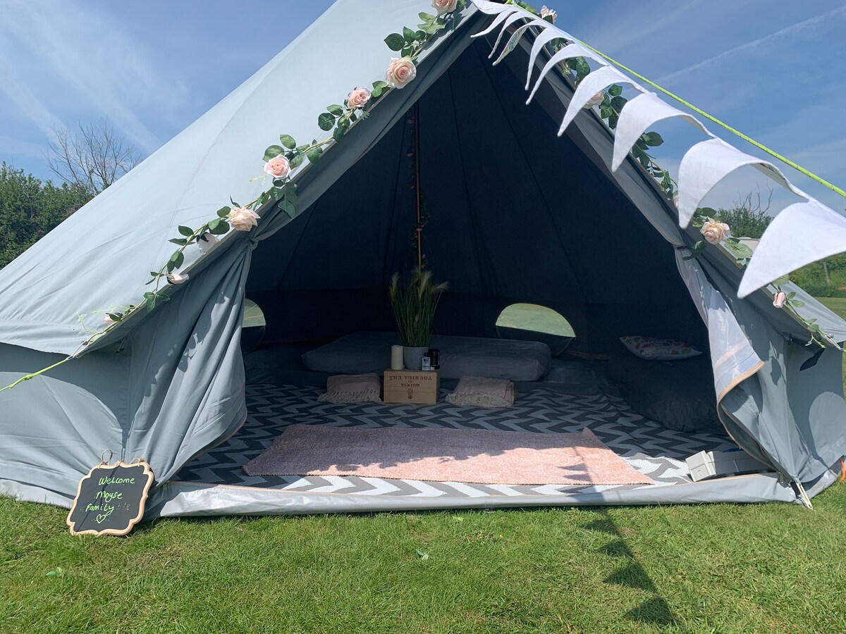 The entrance of a blue bell tent features decorative bunting and floral accents. Inside, a patterned rug covers the floor, while spacious bedding is arranged in a cozy manner. Natural light filters in, creating an inviting space that accommodates multiple guests.