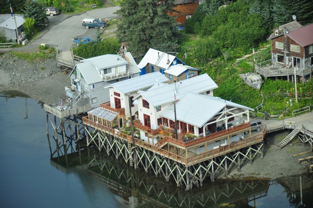 The image captures an aerial view of a timber frame structure elevated on pilings over a tidal estuary. The building features a spacious deck, multiple large windows, and a combination of white and red accents, surrounded by greenery and water.