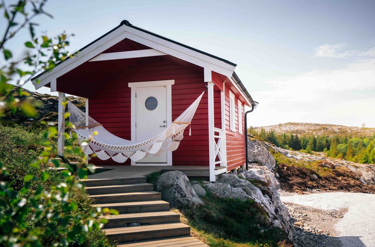 A cozy red cabin is shown with a white door and circular window, nestled among rocky terrain. A hammock is suspended from its porch, inviting relaxation. The surrounding landscape features a mix of greenery and rocks, under a clear sky.