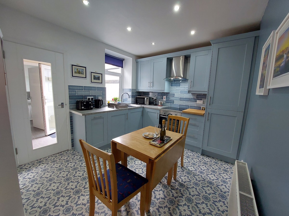 A modern kitchen features blue cabinetry and patterned tile flooring. A wooden dining table is set for two, with a small plate and utensils. Stainless steel appliances and a window provide ample light, while framed artwork adds a personal touch to the space.