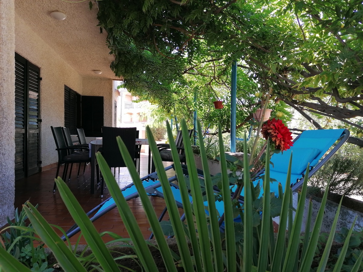 A shaded terrace is shown, furnished with a blue lounge chair and a dining set with chairs. Lush greenery surrounds the area, including potted plants and flowering foliage. Sunlight filters through the leaves, creating a tranquil outdoor ambiance.