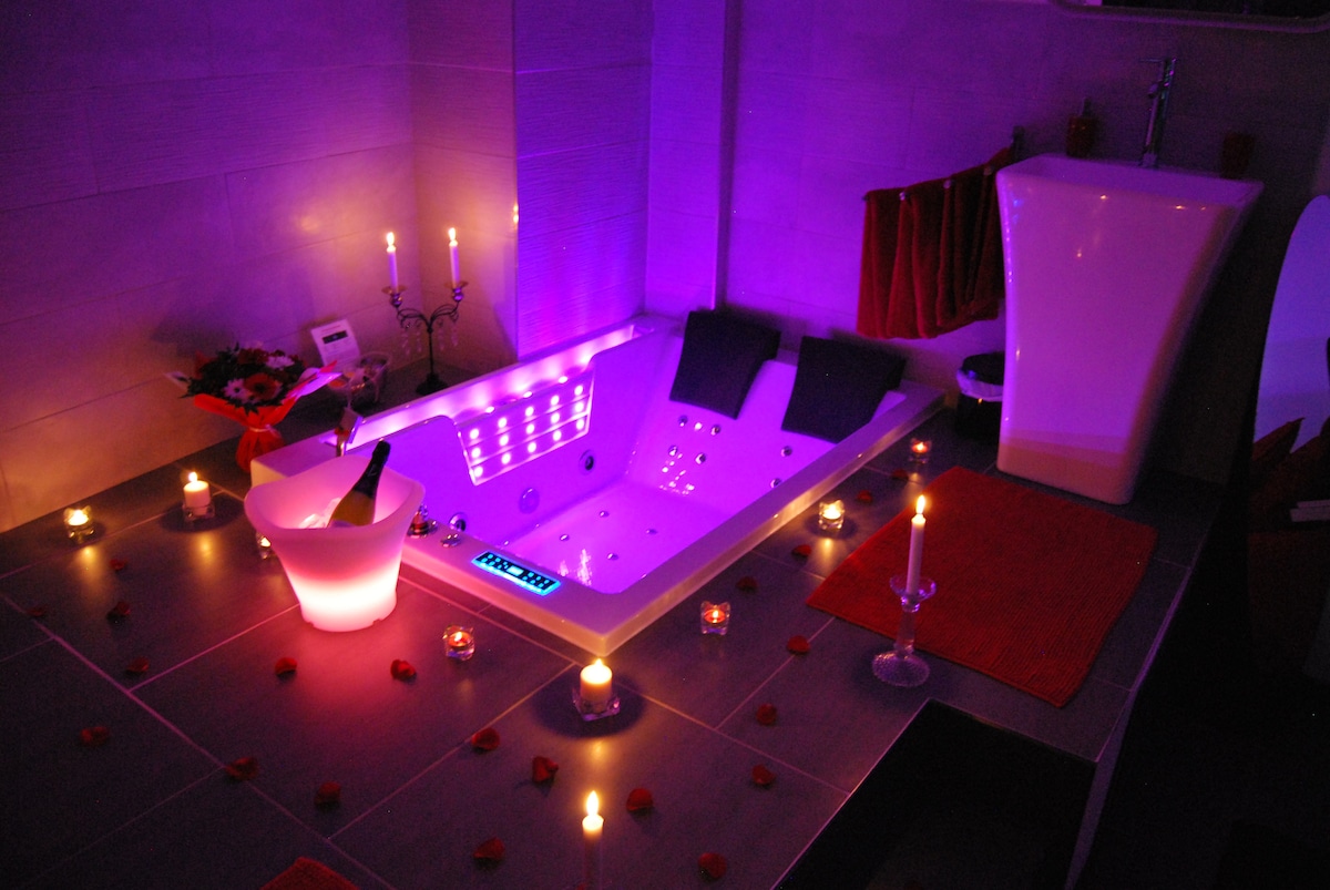 A modern bathroom features a spacious whirlpool bathtub illuminated by soft purple lighting. Surrounding the tub, candles cast a warm glow. Rose petals decorate the surface, while a sleek countertop holds a champagne bucket. A stylish toilet stands nearby, completing the refined aesthetic.