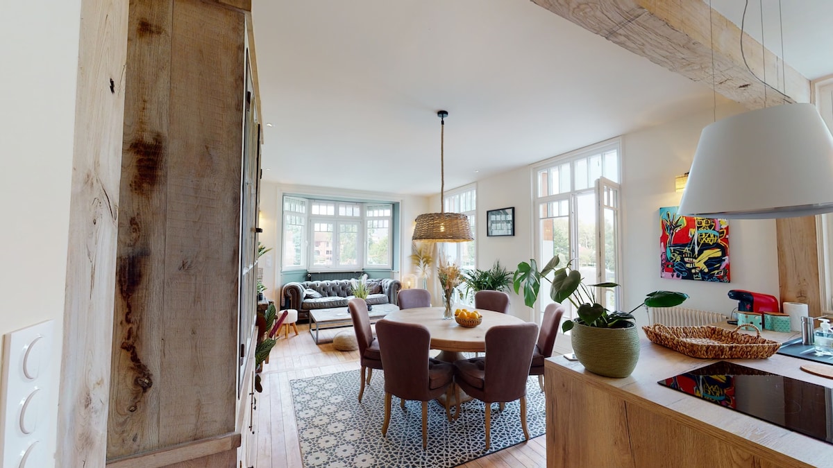 A spacious living area is displayed, featuring a round dining table surrounded by upholstered chairs. A comfortable sofa is positioned near large windows, allowing natural light to fill the room. Decorative plants enhance the inviting atmosphere, complemented by a vibrant piece of artwork on the wall.