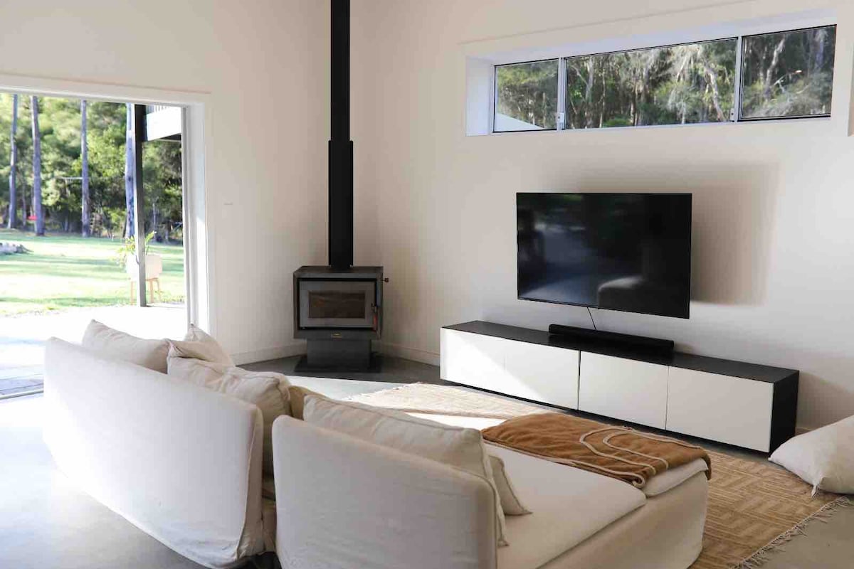 An open living area features a modern sofa positioned near a wood-burning stove. A large television is mounted on a sleek entertainment unit. Natural light enters through expansive windows, creating a bright and inviting atmosphere while overlooking the surrounding nature.