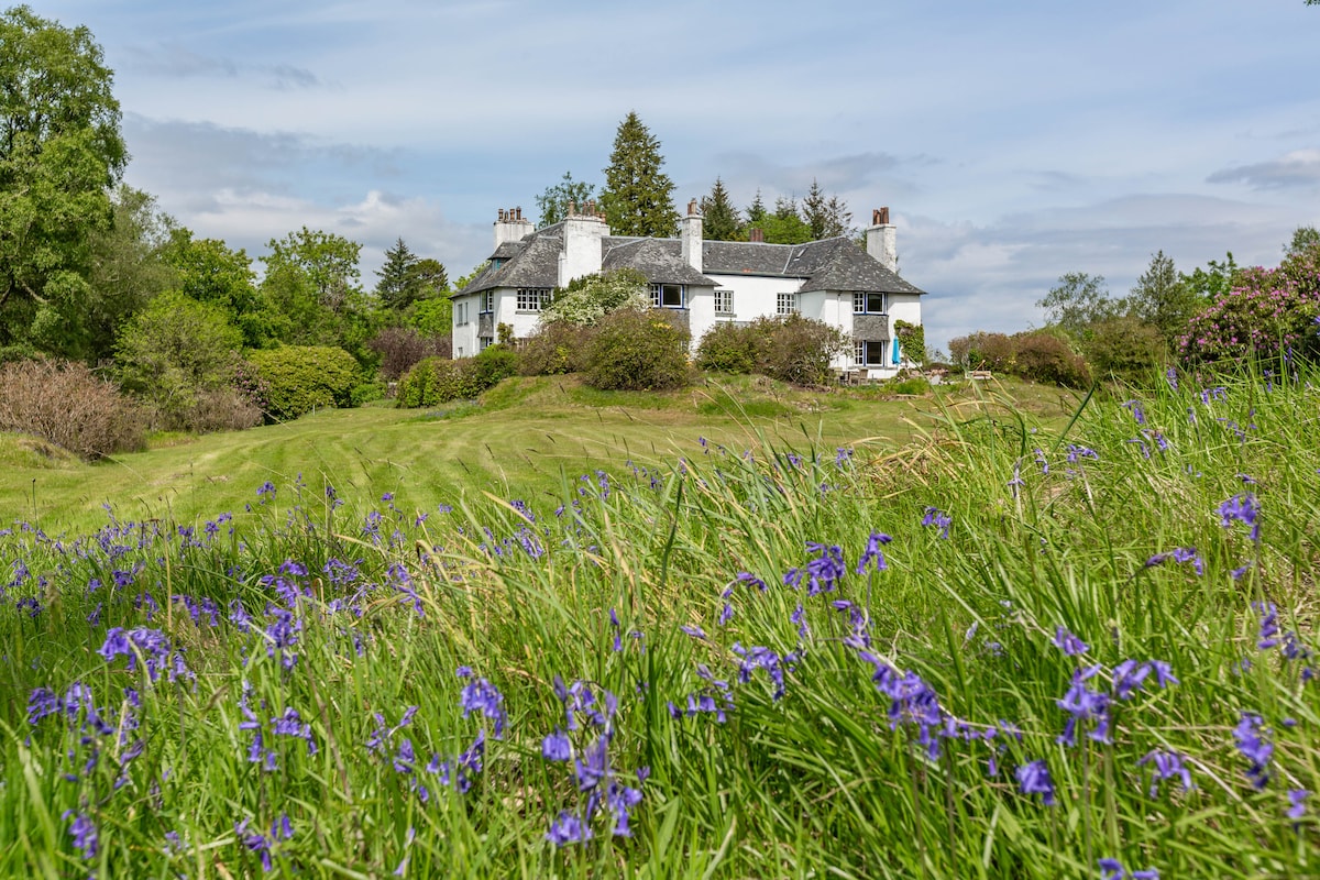 A serene landscape features a traditional white house set amidst lush green grass and blooming bluebells. Rolling hills and verdant foliage create a scenic backdrop, highlighting the home's peaceful location within the natural surroundings.
