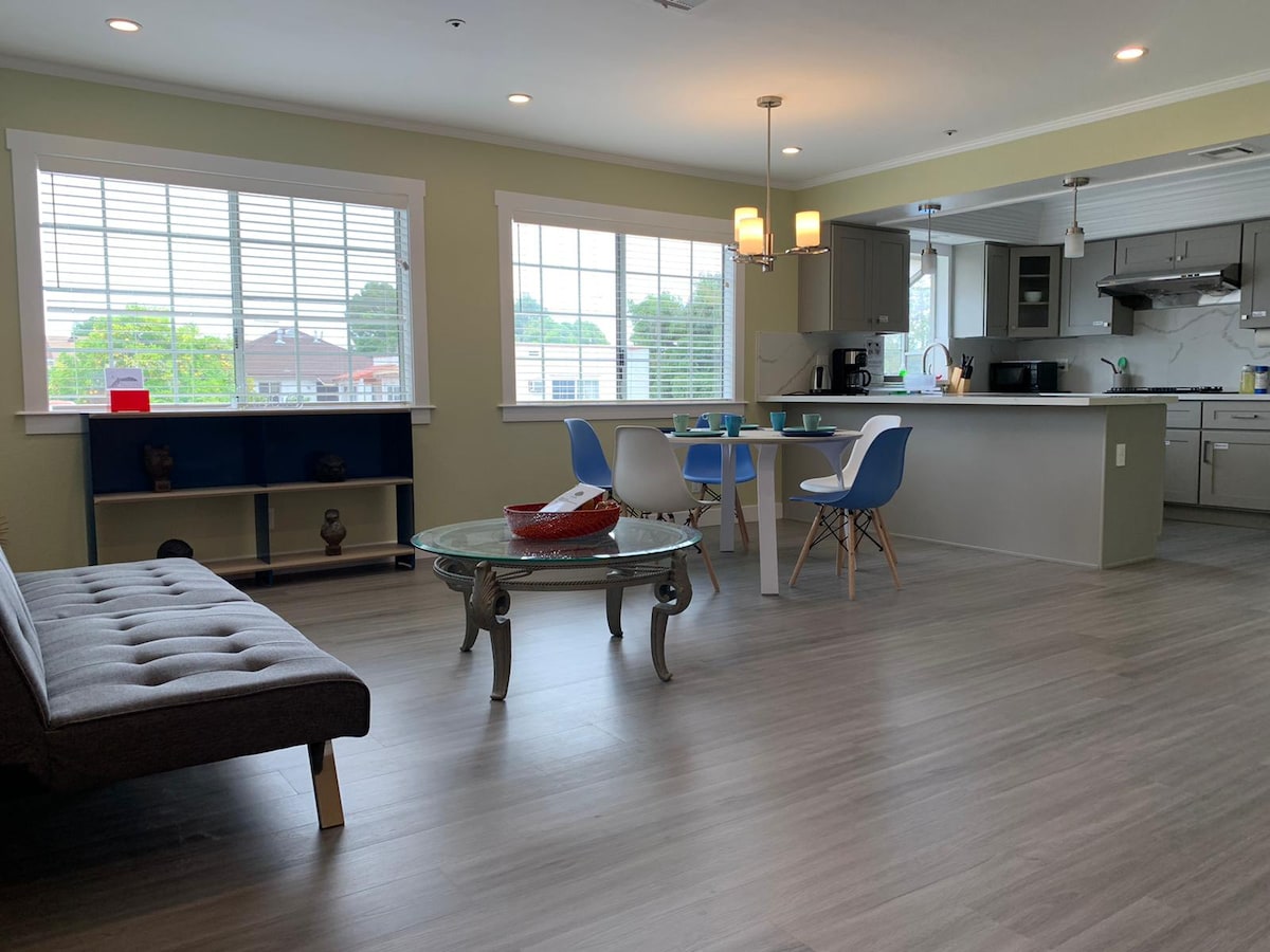 A modern living space features an open floor plan with a stylish gray sofa facing a glass coffee table. A dining area is set with blue chairs around a circular table. Natural light fills the room through large windows with horizontal blinds, illuminating the contemporary kitchen in the background.