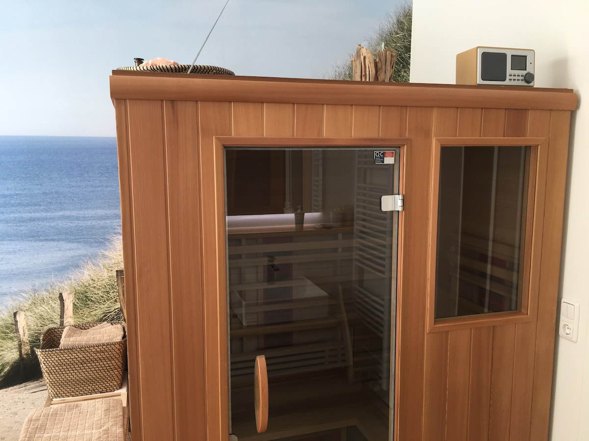 A wooden infrared sauna is pictured, featuring a large glass door allowing visibility of the interior. Soft lighting enhances the natural wood finish, while a small clock and radio are mounted on the wall. The background displays a serene coastal scene.