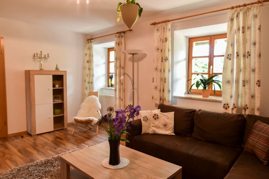 A living area is presented with a brown sofa adorned with decorative cushions. A small coffee table displays a vase of flowers at its center. Natural light enters through the two windows, which are framed by patterned curtains, contributing to a cozy ambiance.
