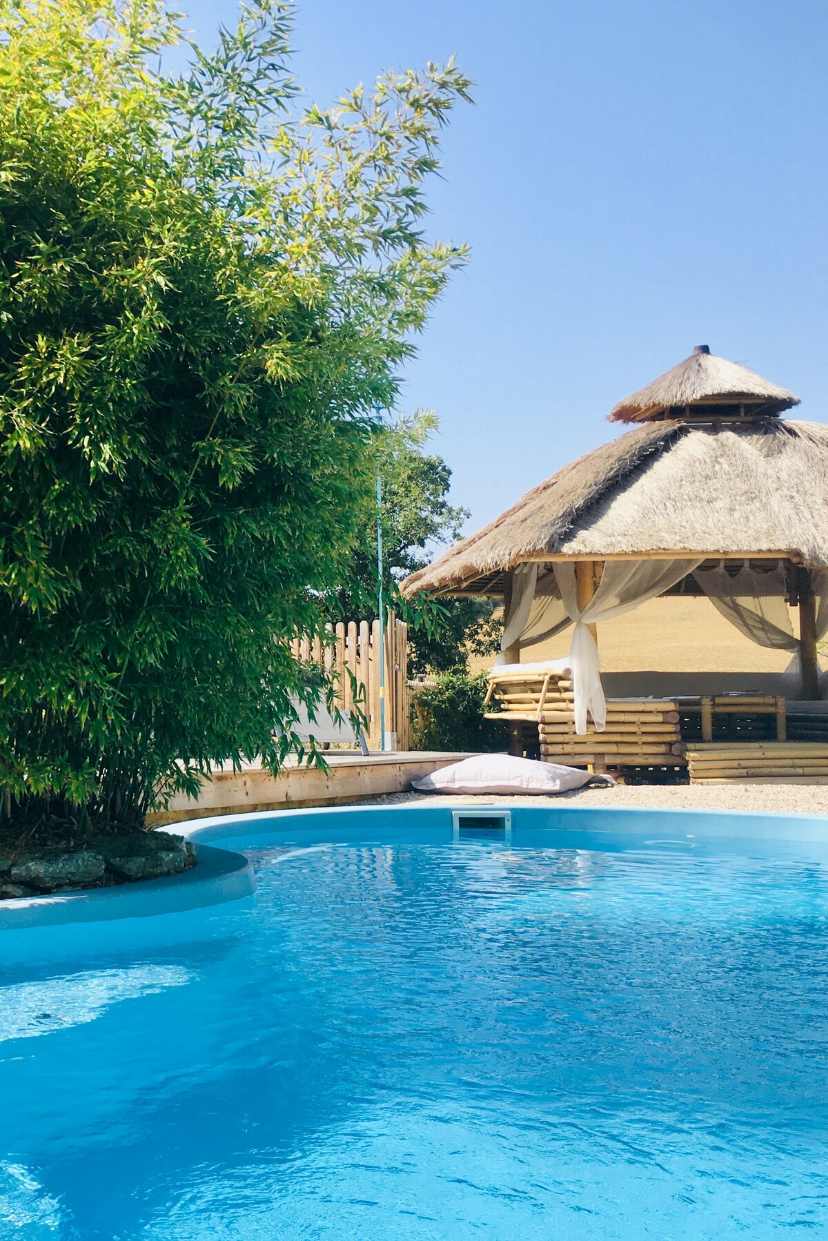 A tranquil pool area is featured, showcasing a shimmering blue pool adjacent to a bamboo tree. A thatched-roof cabana provides shaded seating, enhancing the outdoor relaxation space. Clear blue skies are visible above, contributing to a serene atmosphere.