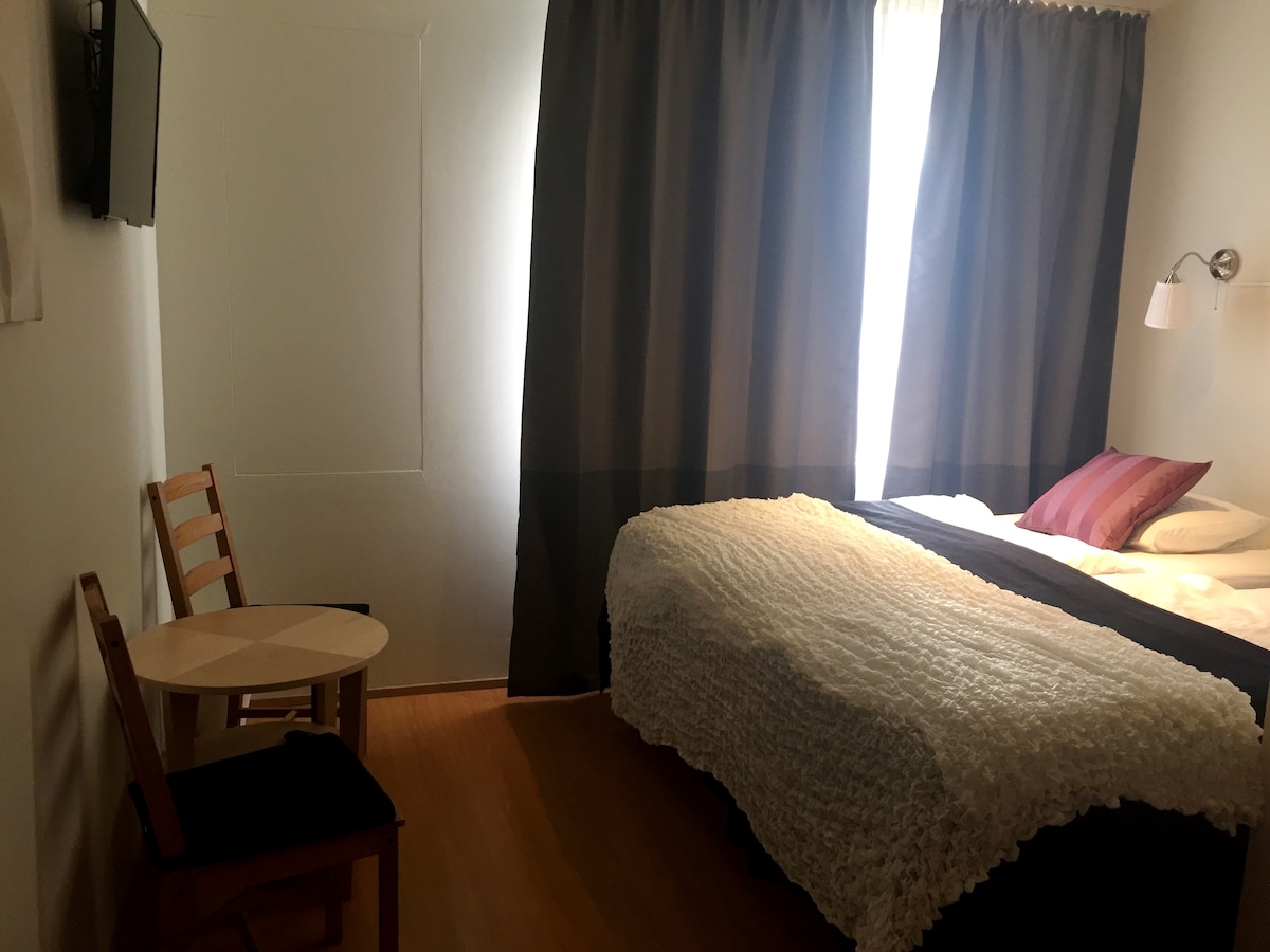 A cozy room is furnished with a spacious bed featuring neutral bedding and accent pillows. A small round table with a wooden chair is positioned near the window, which is covered by dark curtains, allowing natural light to filter into the space.