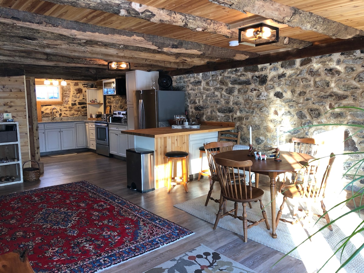 A warm, rustic kitchen and dining area features exposed wood beams and stone walls. Natural light fills the space from a nearby window, highlighting a wooden dining table surrounded by four chairs. A cozy area rug adds comfort to the floor, while a kitchen island offers additional seating.