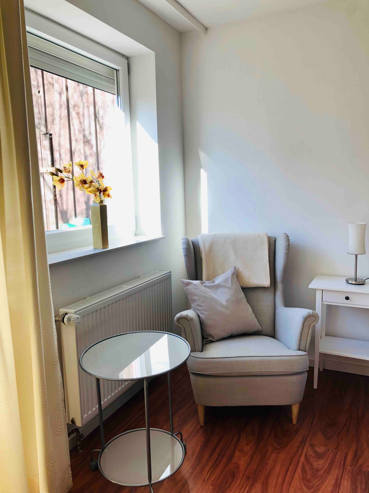 A cozy corner is highlighted by a comfortable armchair with a soft throw and a decorative pillow. A small, round side table is positioned nearby, complemented by a modern lamp. Natural light enters through the window, creating a warm atmosphere.