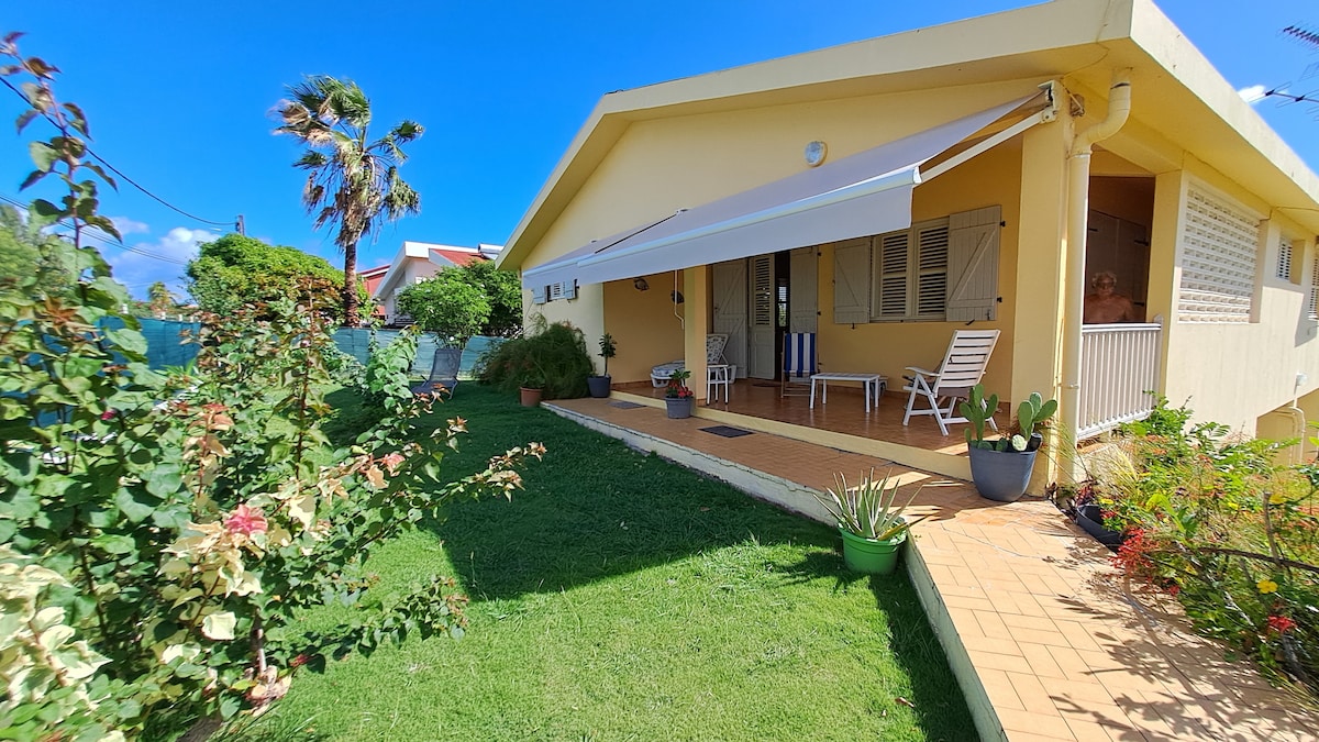 The exterior view of the house highlights a spacious patio area with white sunshades partially extended. Lush green lawn and colorful plants decorate the front yard. A few outdoor chairs are placed on the tiled floor, creating a welcoming entrance.