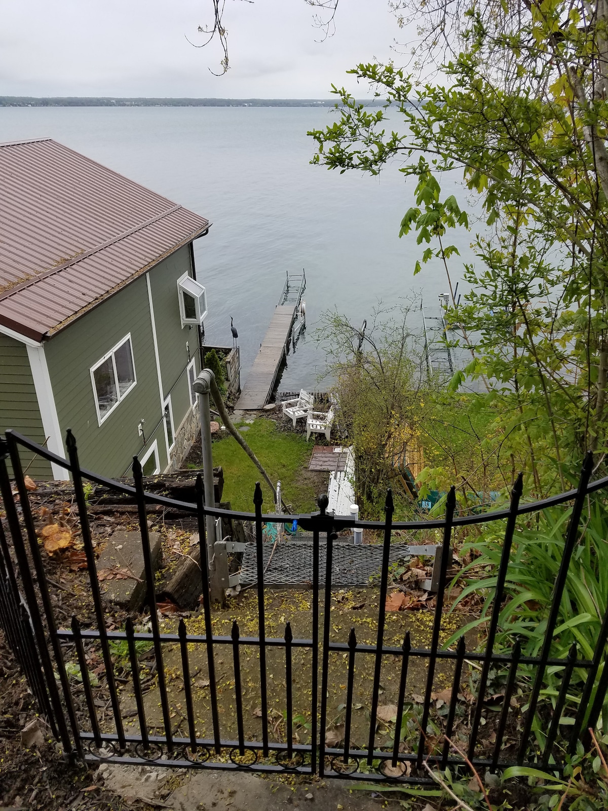 A wrought iron gate leads down a stone pathway towards the water. The green house is visible on the left, with a wooden dock extending into Seneca Lake. Relaxing seating areas and a sandy swim area are seen near the water's edge, surrounded by foliage.