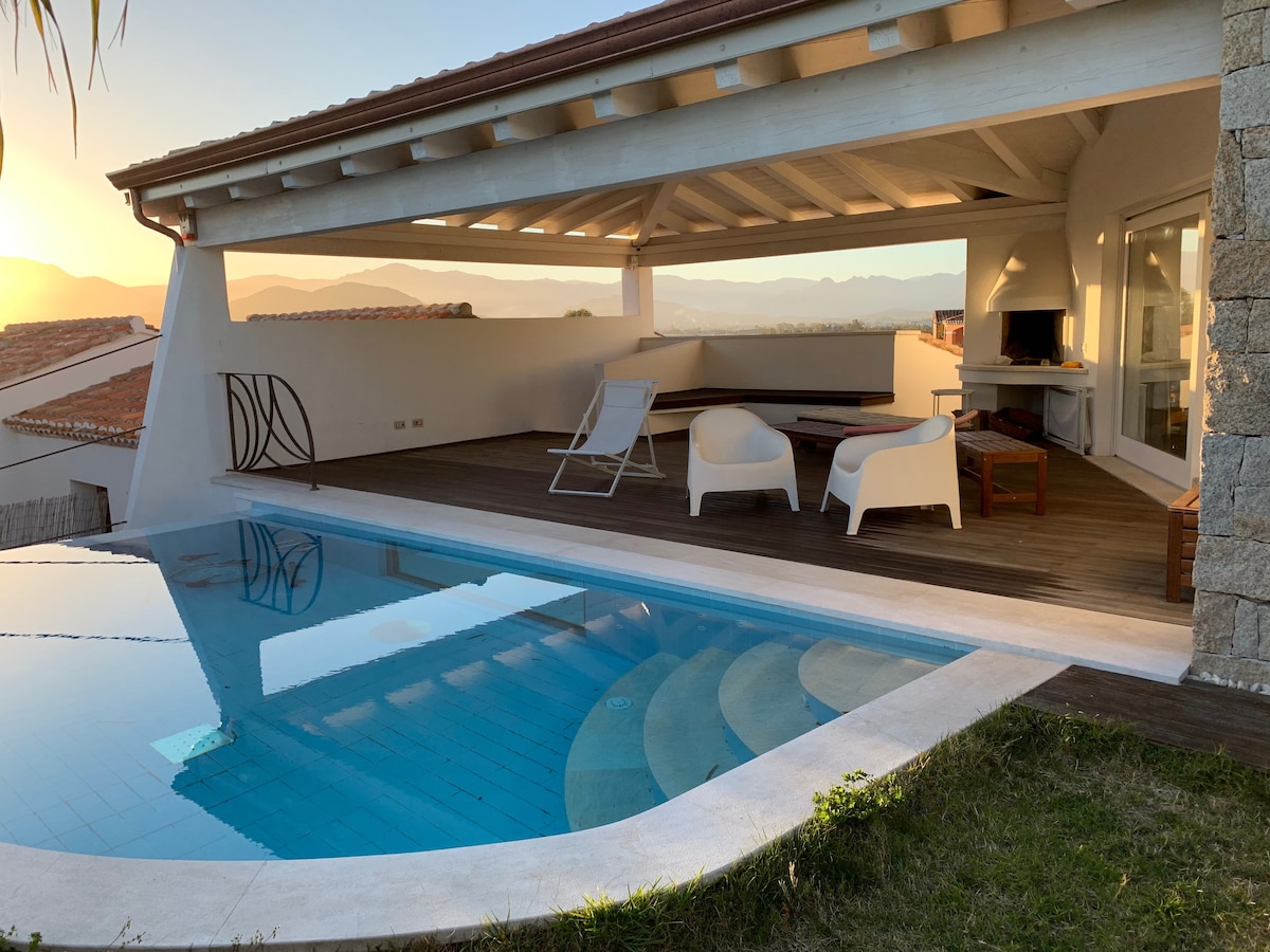 A private pool is visible in the foreground, reflecting the surrounding landscape. A covered patio provides shade with modern lounge chairs arranged nearby. A cozy seating area is also present, enhancing the outdoor space. The backdrop features distant mountains and a clear sky.