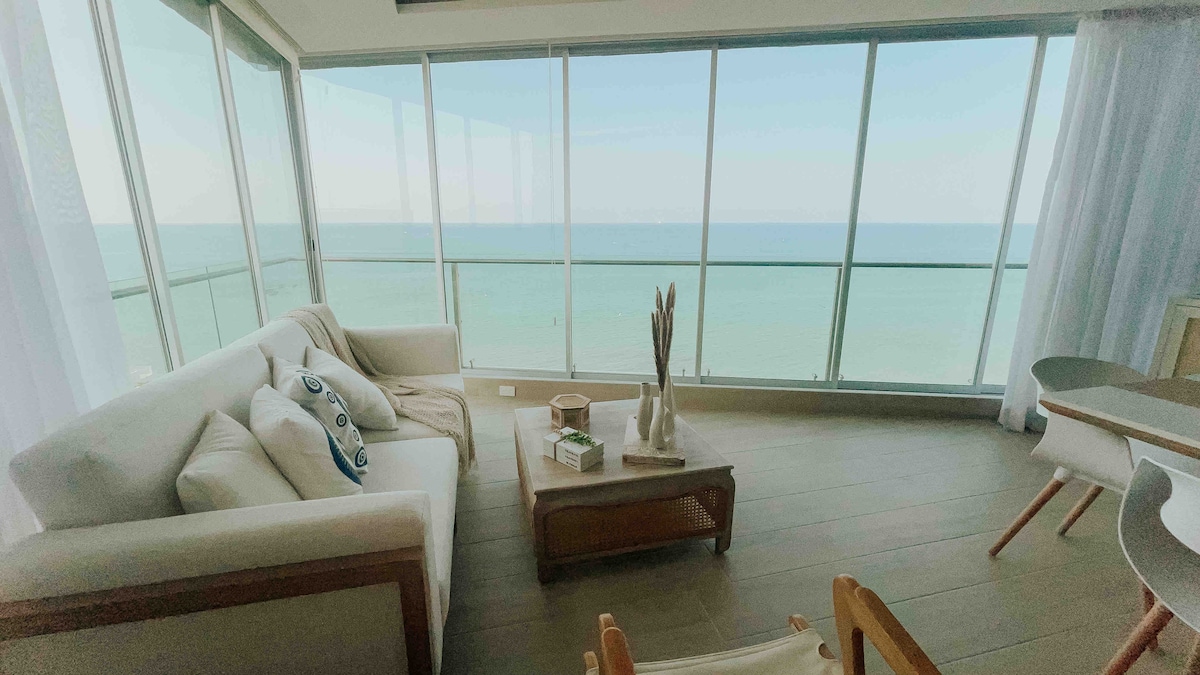 A bright living area features large glass windows offering a clear view of the ocean. A comfortable white sofa is placed alongside a wooden coffee table, which holds decorative items. Light, airy curtains gently frame the space, enhancing the coastal ambiance.