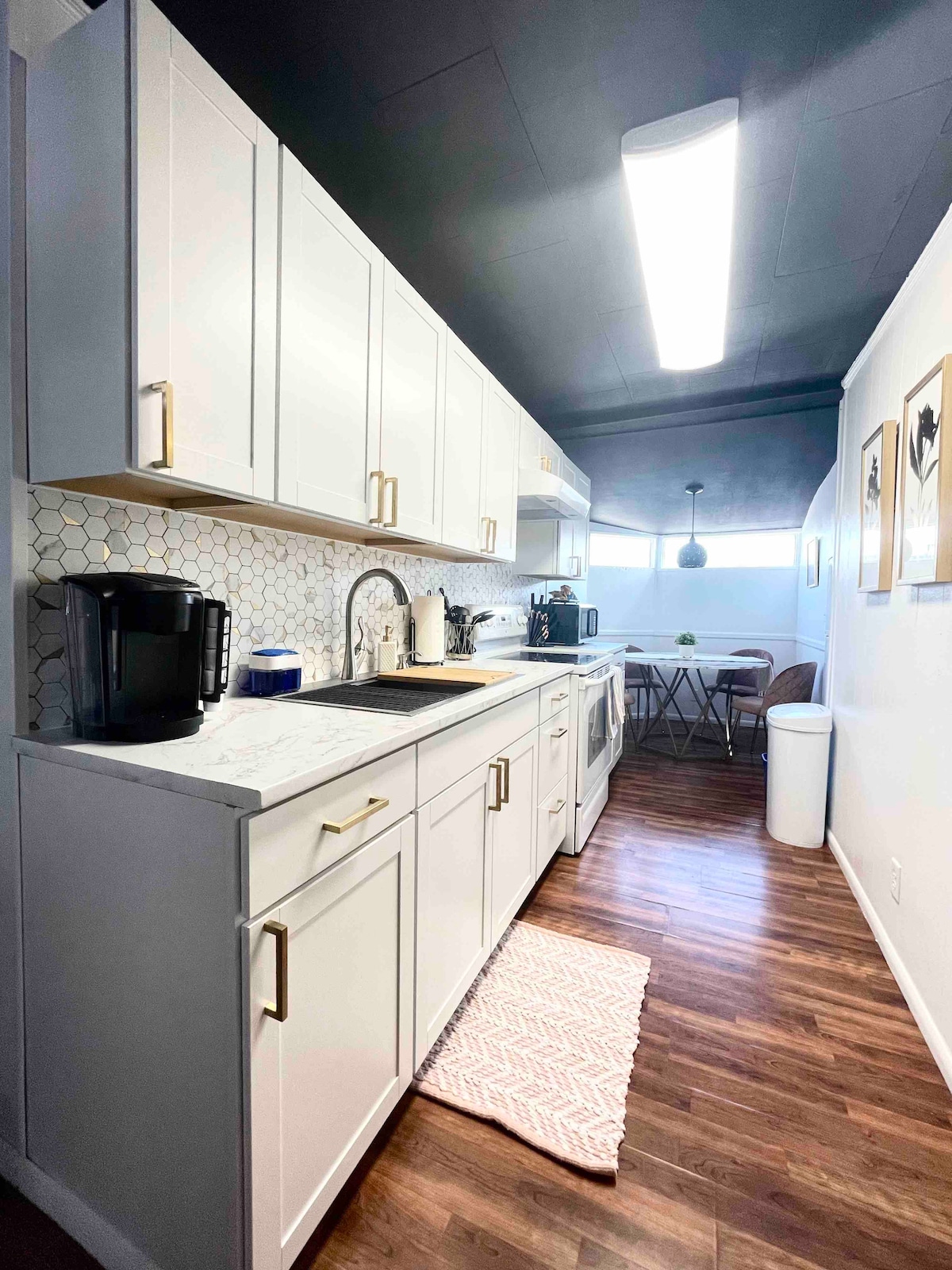 A modern kitchen features sleek white cabinetry with gold hardware. A countertops houses a coffee maker and other appliances. The space is complemented by a subtle hexagonal tile backsplash and a small dining area with a round table and chairs, illuminated by natural light.