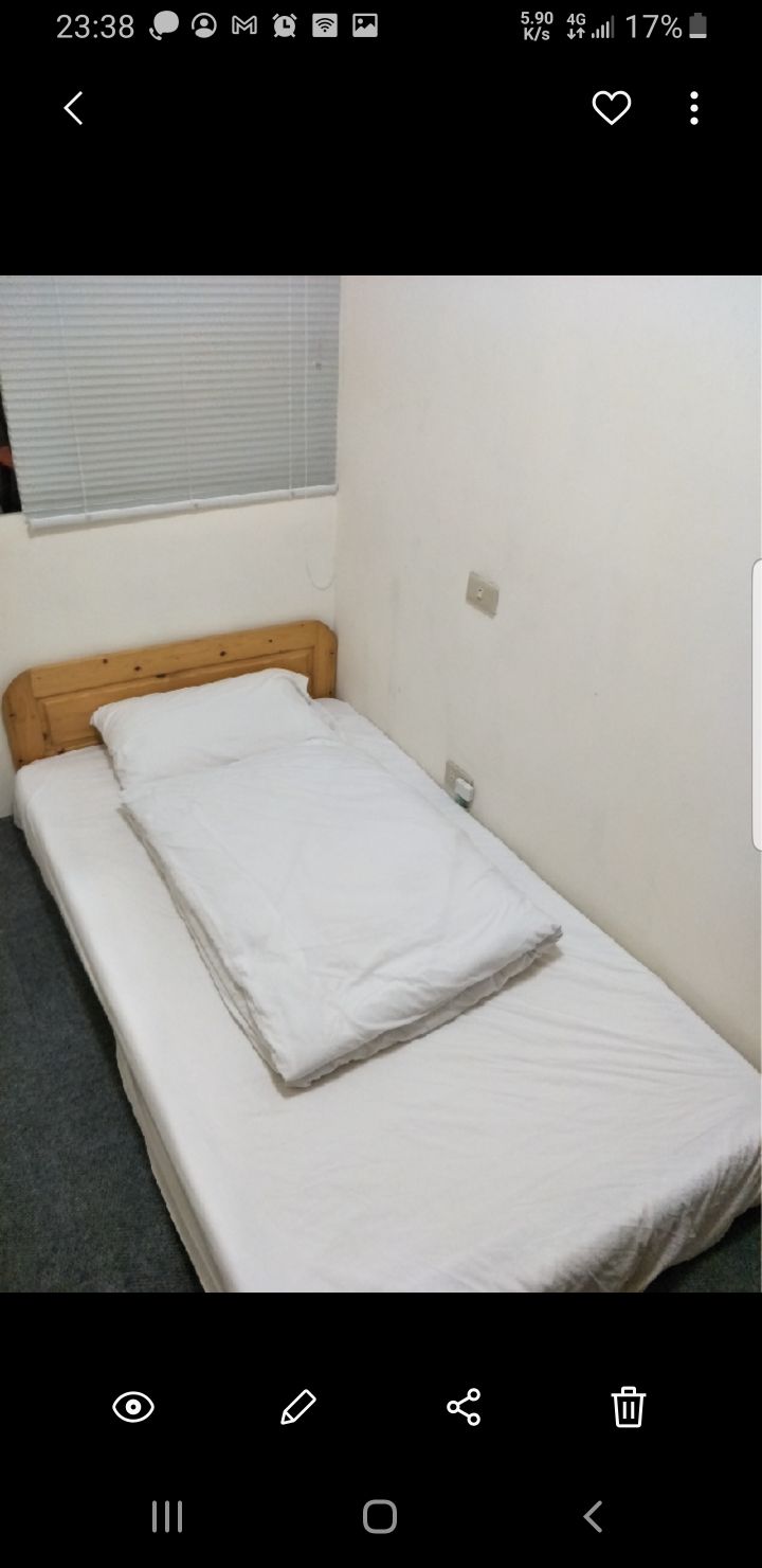 A simple sleeping area is shown with a wooden bed frame and a neatly made mattress. A white blanket is laid over the bed, while a single pillow rests against the headboard. A window with blinds allows natural light to enter the space.