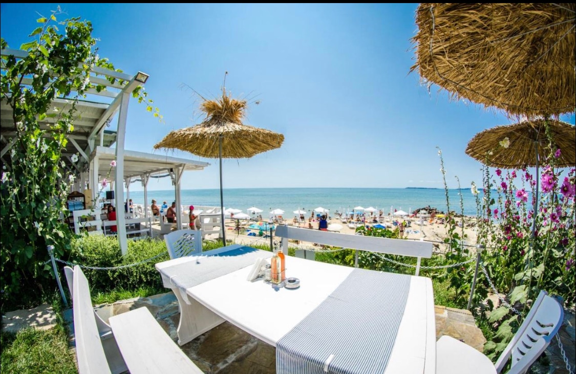 A dining area is set outside, featuring a white table and chairs under a straw umbrella. The view extends to a serene beach with umbrellas and sun loungers. Lush greenery surrounds the area, while the ocean glistens under clear blue skies.