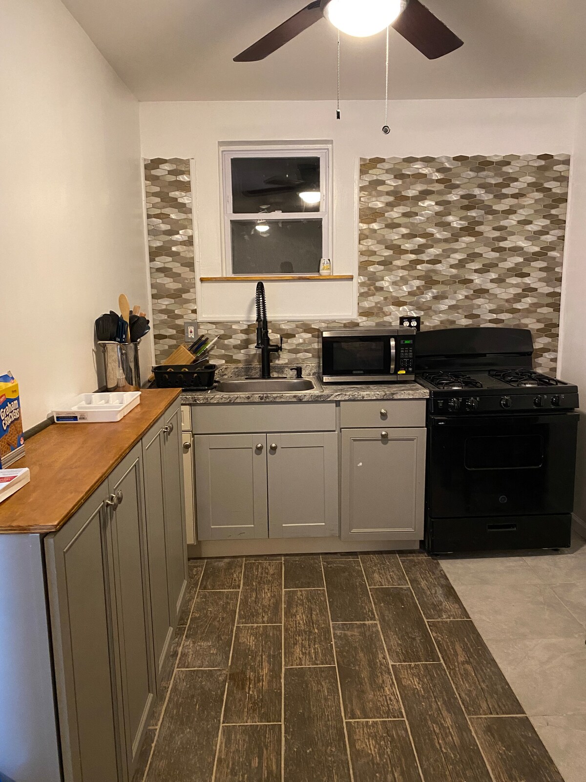 A compact kitchen area is equipped with essential appliances, including a microwave and a gas stove. Modern cabinetry complements a unique backsplash, while a window provides natural light. A wooden countertop offers additional workspace, with utensils and cooking tools neatly arranged.