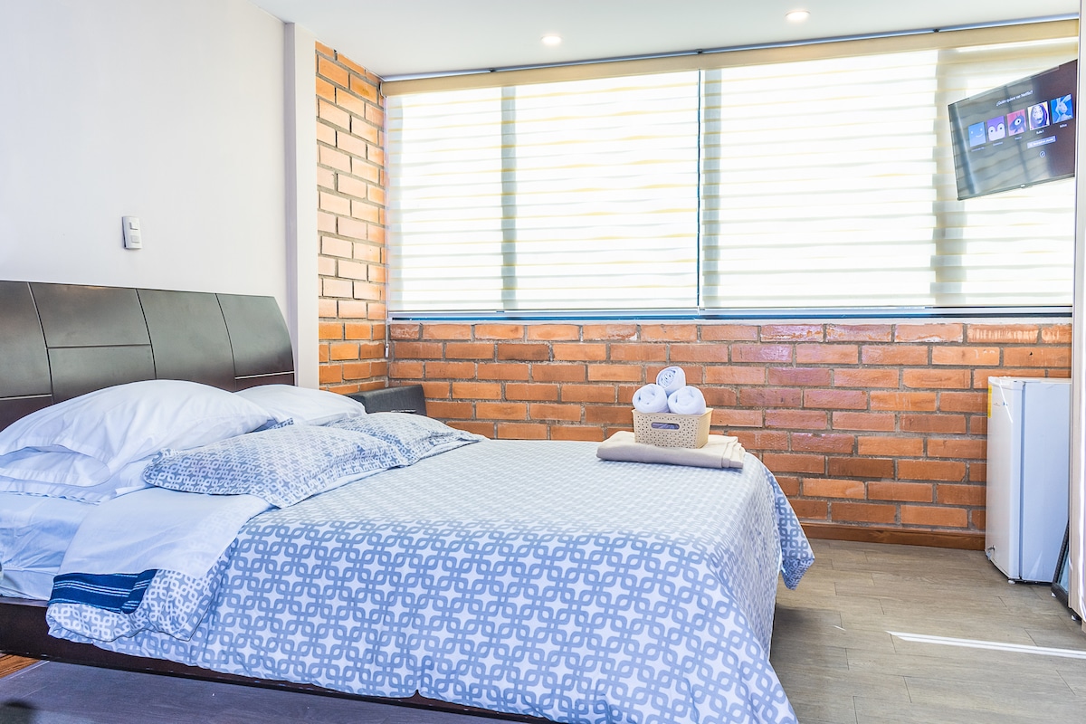 A modern bedroom features a spacious bed with crisp white linens and a patterned blanket. Soft natural light filters through the window with layered blinds, illuminating the exposed brick wall. A small TV is mounted on the wall, and a basket of towels is placed on the bed.