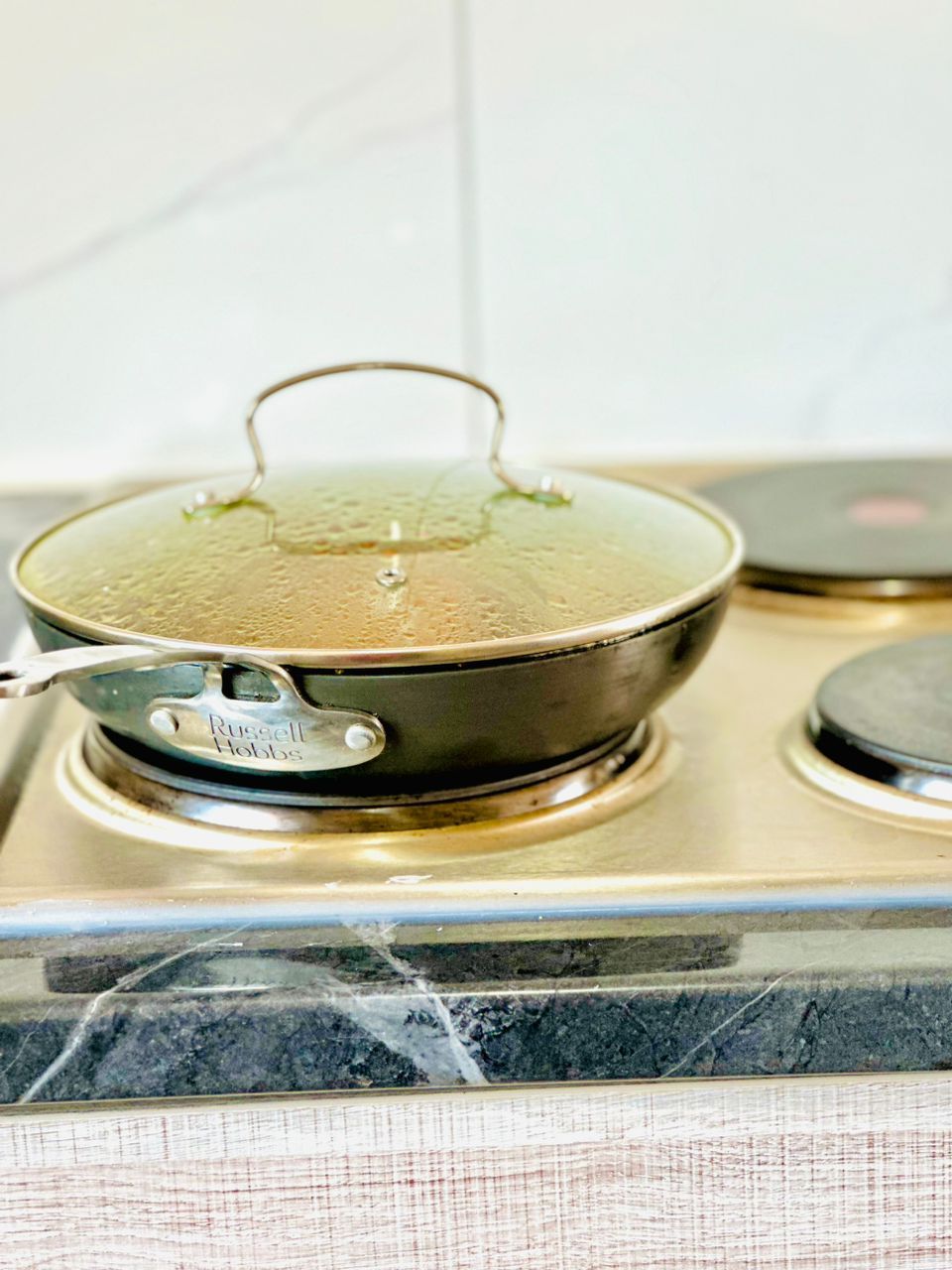 A sleek cooking pan with a glass lid is positioned on a shiny stove. Condensation can be seen on the lid, suggesting food is being cooked. The stovetop features two additional burners, each with a circular surface. A smooth, dark countertop provides a stylish backdrop.