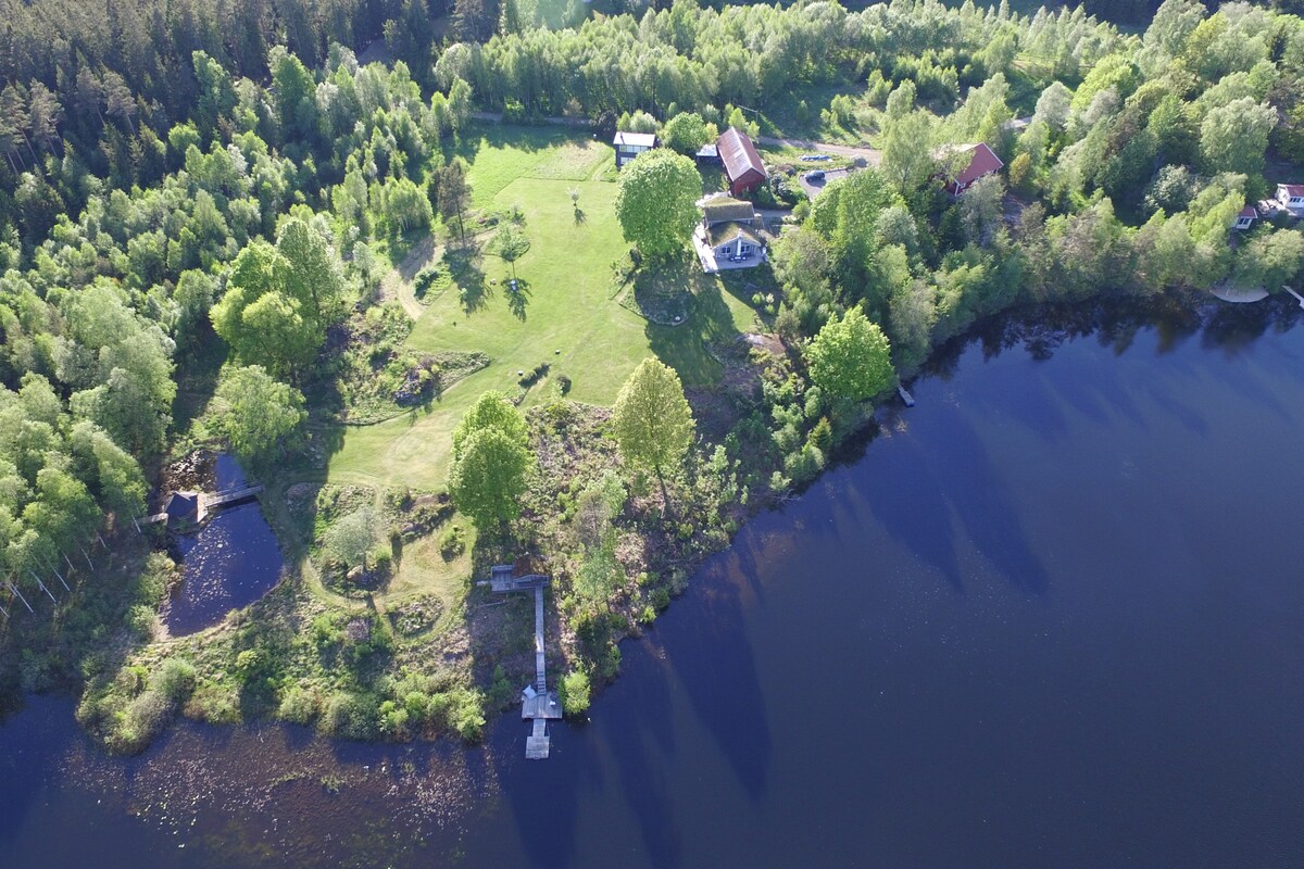 An aerial view captures a sprawling green landscape bordered by a large lake. The property features a mix of trees and open spaces, with various structures visible, offering a serene retreat surrounded by nature.