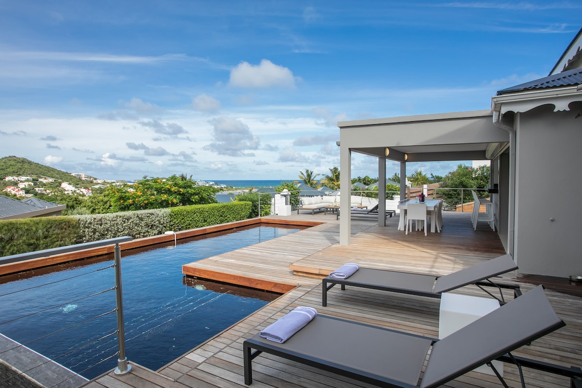 An outdoor patio area features sleek sun loungers and a wooden deck surrounding an infinity pool. A dining table is positioned nearby, with scenic views of the bay and lush greenery visible in the distance under a partly cloudy sky.