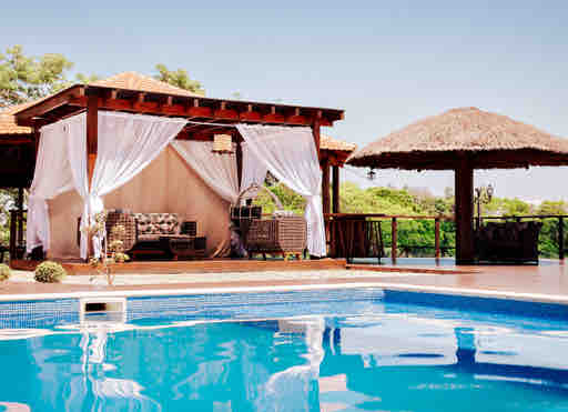 A shaded gazebo constructed with wooden beams and a thatched roof is situated near a sparkling outdoor pool. Comfortable seating is provided under flowing white curtains, while lush greenery surrounds the space, creating a peaceful atmosphere for relaxation.