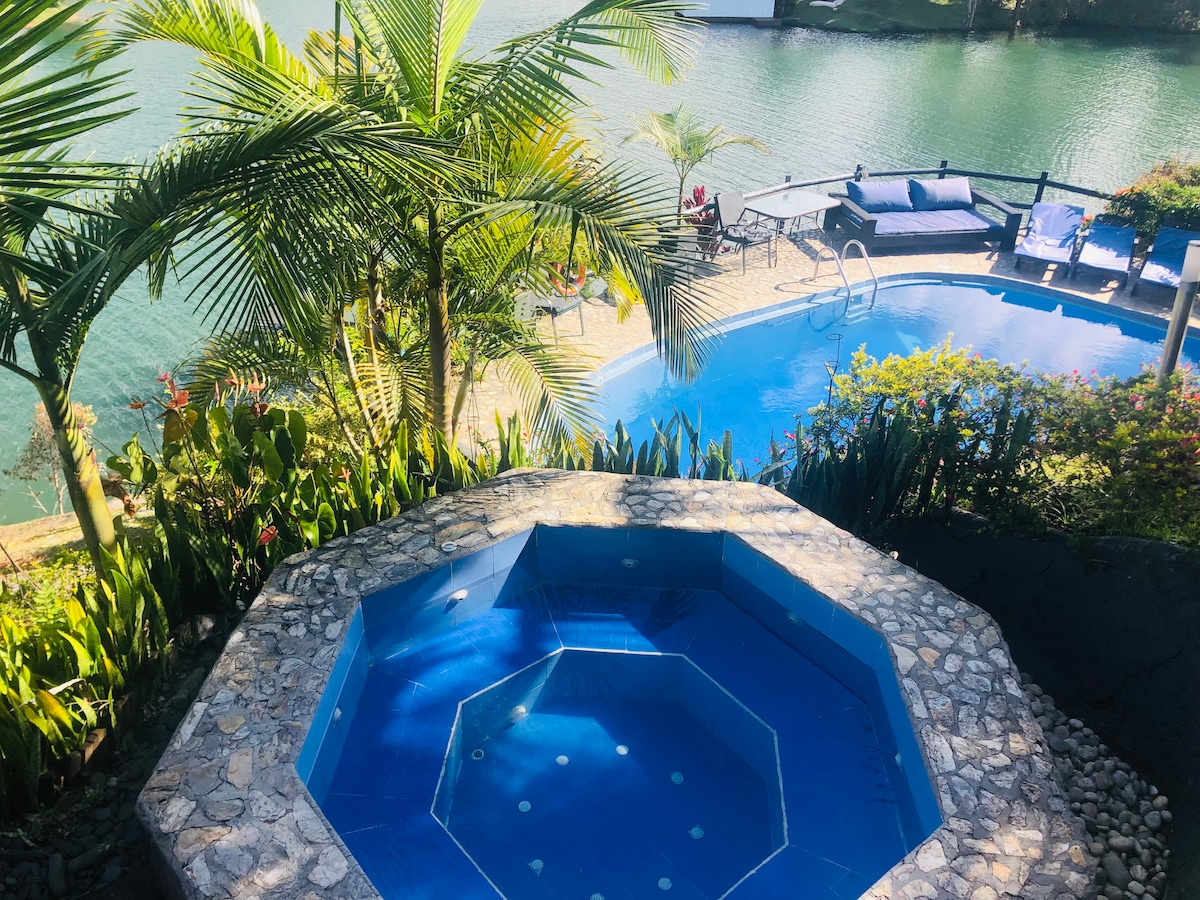 A hot jacuzzi with a stone border is set on a terrace overlooking a serene lake. In the background, a spacious pool with lounge chairs and tropical plants can be seen, enhancing the tranquil atmosphere. The lush greenery surrounds the area, creating a private retreat.