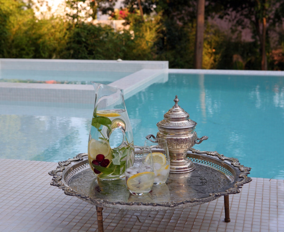 A silver tray sits on a surface near the swimming pool, showcasing a glass pitcher filled with refreshing drink garnished with herbs and fruits. Two glasses, also containing the drink, rest on the tray beside an ornate silver container, reflecting the serene blue waters of the pool.