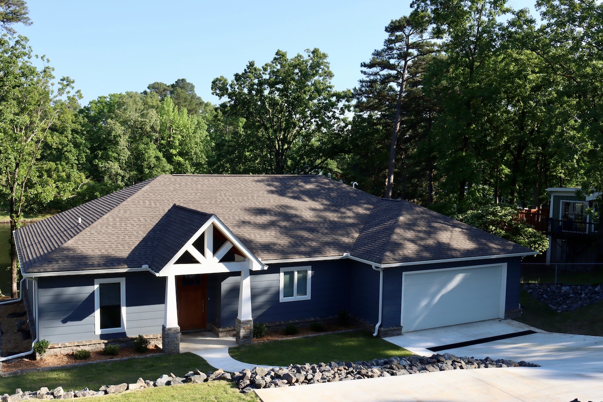 The exterior of a modern lakefront house is shown, featuring a mix of dark siding and stone accents. A large driveway leads to an attached garage, surrounded by a well-kept yard with lush greenery and natural landscaping. Trees provide shade, enhancing the serene environment.