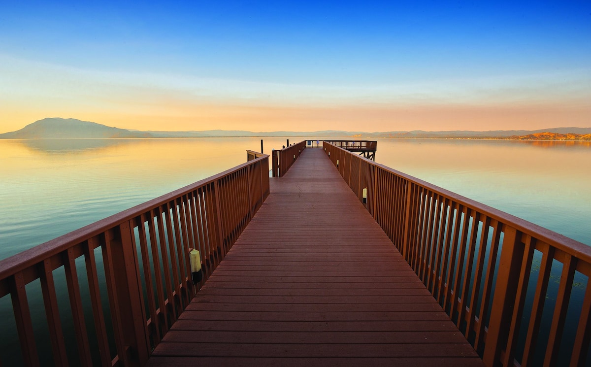 A wooden pier extends into a tranquil lake, surrounded by calm waters that reflect the warm colors of the sky. The view includes gentle mountains in the background, creating a serene and expansive atmosphere.